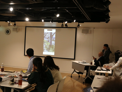 室内で参加者たちが前方スクリーンに映された屋外風景の映像に注目しながら説明を聞いている写真