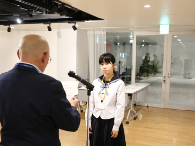 男性がマイク越しに女子生徒へ文書を読み上げ、女子生徒が正面で緊張した面持ちで受け答えの姿勢を取っている写真