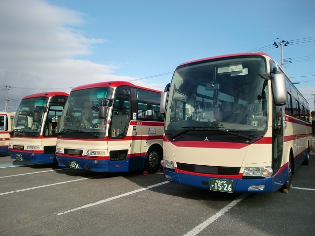 駐車場に一列に並んで停車している、クリーム色の車体に、赤のラインが入り、下部が青色に塗り分けられた三台の大型バスを斜め前から捉えた写真