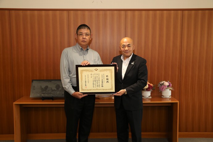 木目調の壁を背景に、グレーのシャツを着た男性と大寺市長が並んで立ち、額に入った感謝状を正面に向けて掲げている贈呈式の記念写真