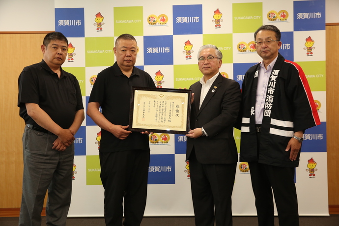 須賀川市のロゴやキャラクターが描かれたバックパネルの前で左から中道利高専務取締役、中道史和代表取締役社長、橋本市長、五十嵐伸須賀川市消防団団長が並び社長と市長で感謝状を持っている写真
