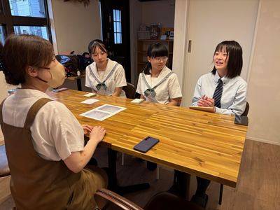 木製テーブルを囲んで3人の女子生徒と女性店員が明るく落ち着いた室内で話し合う様子を撮影した写真