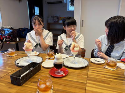 木製テーブルで3人の女子生徒が彩り豊かなパフェや冷たい飲み物を味わいながら穏やかに歓談する様子の光景を写した写真
