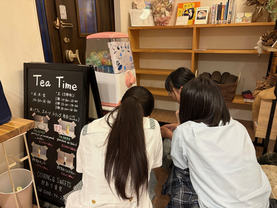 店内の木製棚やガチャ機の前で女子生徒3名が黒板メニューを覗き込み何かを確認している様子を撮影した写真