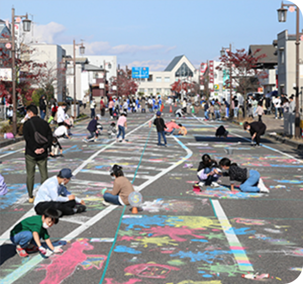 歩行者天国となった道路の路面に、多くの子供や大人がカラフルなチョークで絵を描いている様子。