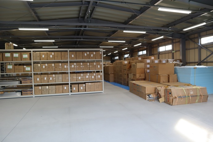 4段のスチール棚に同じ大きさの段ボール箱が整然と並べられ、広い倉庫内には他にも積み上げられた段ボールや梱包材が多数見える様子の写真