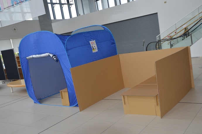 青色のポップアップテントと段ボールのパーテーションが設置されている、災害時の避難空間を想定した展示の様子の写真