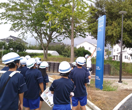 紺色の体操服と白いヘルメットを着用した生徒たちが、屋外で青い縦長の看板を見上げており、ヘルメットをかぶった男性が看板の目盛りを指さしながら説明している様子の写真