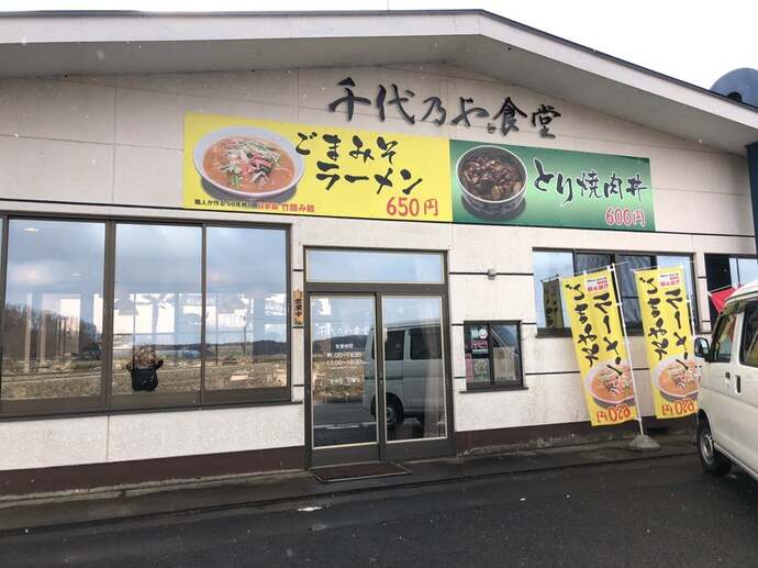 「千代乃や食堂」と書かれた看板の下に「ごまみそラーメン 650円」と「とり焼肉丼 600円」の写真がが掲げられた外観の全体を捉えた写真