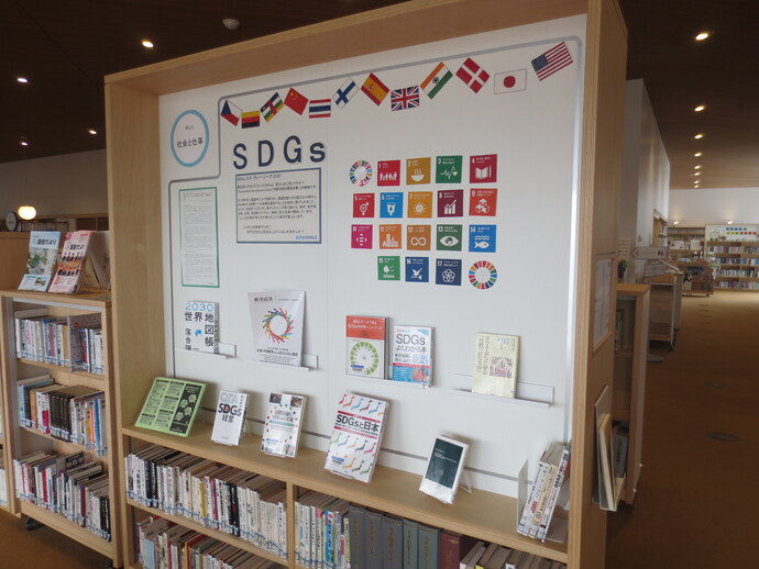 図書館内の木製書棚に設けられたSDGs特設コーナーで、中央の白いパネルには17のカラフルな目標アイコンと説明文、上部には複数の国の国旗が並び、下の棚にはSDGs関連書籍が展示されている写真