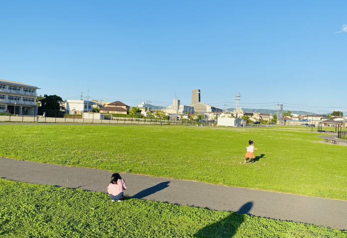 晴天の下、広々とした芝生広場で、手前の歩道に座り込む子どもと奥の芝生を元気に走り回る子どもの姿が見える大黒町防災公園の写真