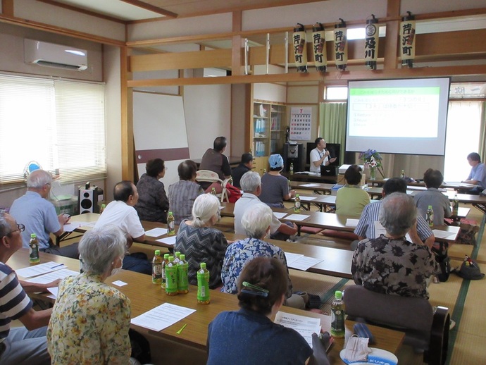 和室で、多くの高齢者が机に向かって座り、前方のスクリーンに映し出されたスライド資料を見ながら関係者の話を熱心に聞いている写真