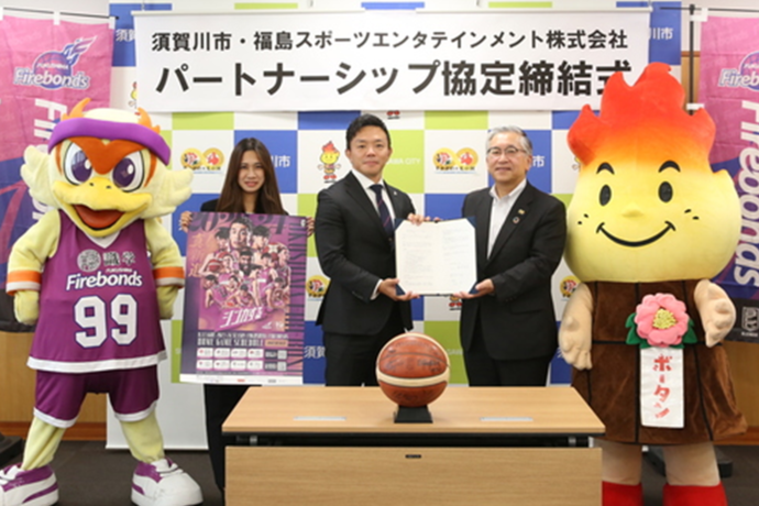 須賀川市と福島スポーツエンタテインメント株式会社のパートナーシップ協定締結式において、スーツ姿の男性と女性と2体のマスコットキャラクターがテーブルの前に立ち中央のスーツ姿の男性2人が書類を一緒に持って記念撮影を行っている様子の写真