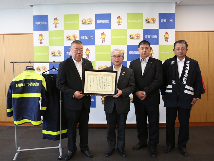 須賀川市のロゴやキャラクターが描かれたバックパネルの前で左から中道史和代表取締役社長、橋本市長、中道利高専務取締役、五十嵐伸須賀川市消防団団長が並び社長と市長で感謝状を持ち、左端には防火服が掛けられている写真