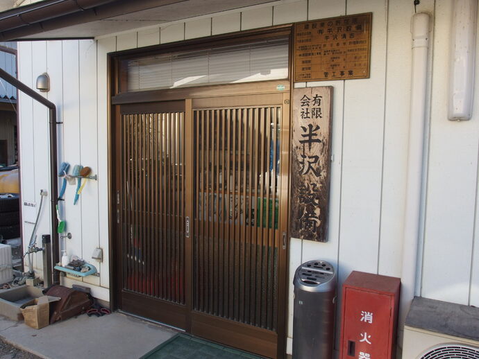 白い壁の建物の両開き玄関の右側に、縦書きで「有限会社半沢設備」と彫られた木製の看板が掲げられている写真