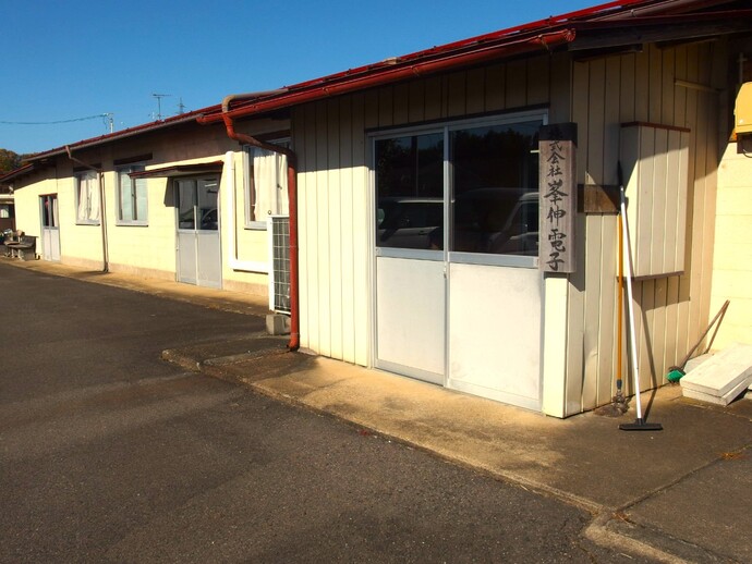 明るい日差しの下、薄い黄色の外壁と茶色の庇を持つ平屋の建物が写っており、掃き出し窓の横には「株式会社峯伸電子」と書かれた立て看板がある外観写真