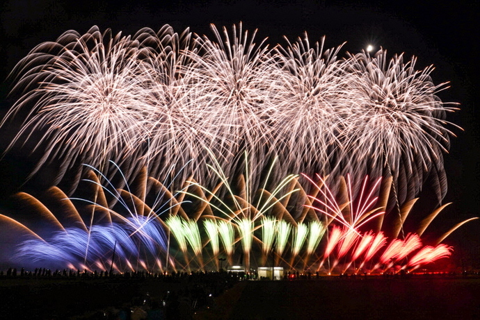 夜空一面に、壮大に広がる白色の打ち上げ花火と、地上近くで扇状に広がる青、緑、赤色の花火が同時に打ち上げられている写真