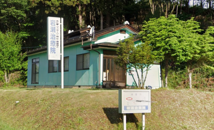 緑の芝生の斜面上に水色系の外壁と茶色の瓦屋根を持つ平屋建ての建物が建ち、その左側に「岩瀬治療院」と書かれた白い看板が設置されている写真