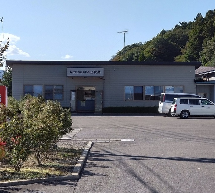 2台の車がどまった駐車場の後ろに、灰色の壁で横長の建物が建ち、入口の上に「株式会社いわせ食品」と看板が出ている株式会社いわせ食品の外観写真