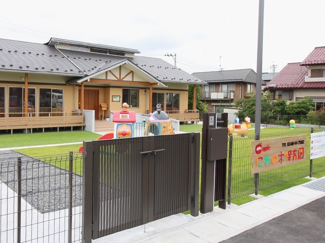 黒い門のついたフェンス囲まれた敷地に、キャラクターがデザインされた遊具が複数設置された芝生の園庭が広がり、奥に平屋建ての園舎がたっている「にれの木保育園」の外観写真