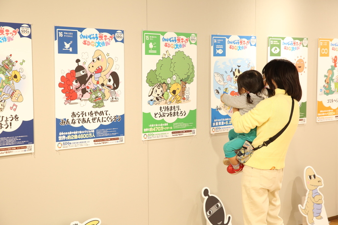「かいじゅうステップSDGs大作戦」と書かれた、様々なイラストが描かれたポスターが壁一面に並んでいるのを幼い子どもを抱えた親子連れが眺めている様子の写真
