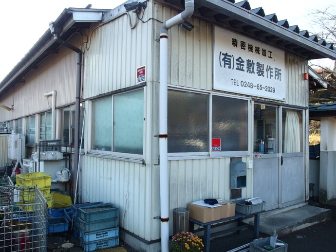 白い外壁で、入り口の上部に「精密機械加工 有限会社金敷製作所」と書かれた看板が掲げられている、有限会社金敷製作所の外観写真