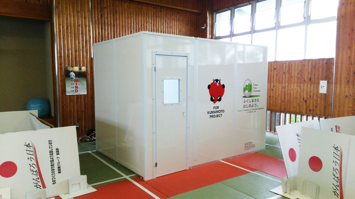 木目調の壁の広い屋内スペースの中央に、熊本支援プロジェクトのロゴやメッセージが描かれた白いパネル製の仮設個室が設置され、周囲には「がんばろう日本」と書かれた白いボードが置かれている写真