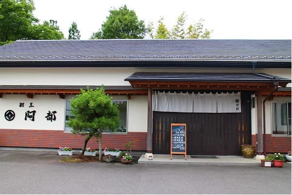 白壁と茶色の瓦屋根の日本家屋風の建物で、左側の壁に「阿部」の文字が描かれ、中央の入り口に白色の暖簾と黒いメニューボードが設置されている外観写真