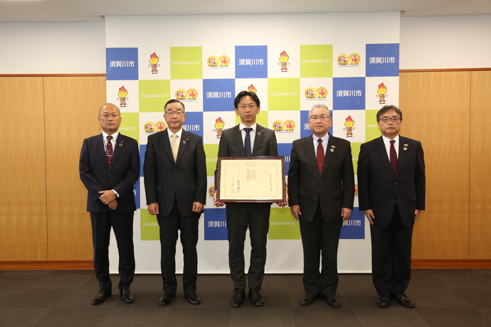 市のロゴが並ぶ背景の前で五人のスーツ姿の人物が大きな賞状を中心に端正な姿勢で整然と立ち並ぶ紺綬褒章伝達式の写真