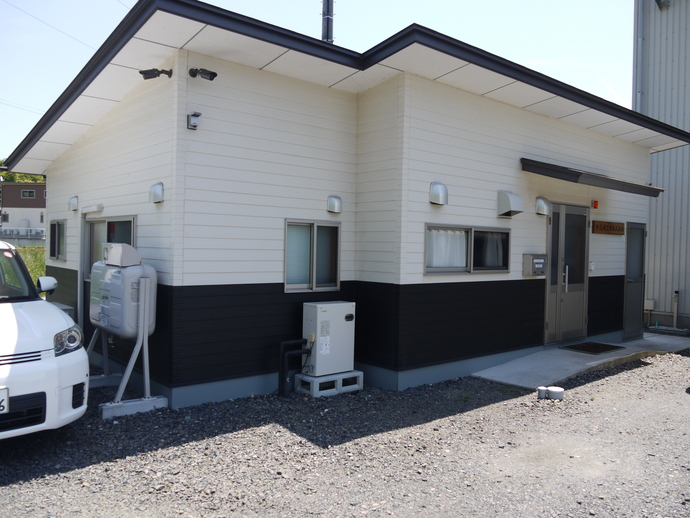 黒と白の横張りサイディングで仕上げられた平屋の建物が、砂利の敷地内に建っており、正面の入り口付近には給湯器のような室外機と、左側には白い軽自動車が一部写り込んでいる今設備工業株式会社の外観写真