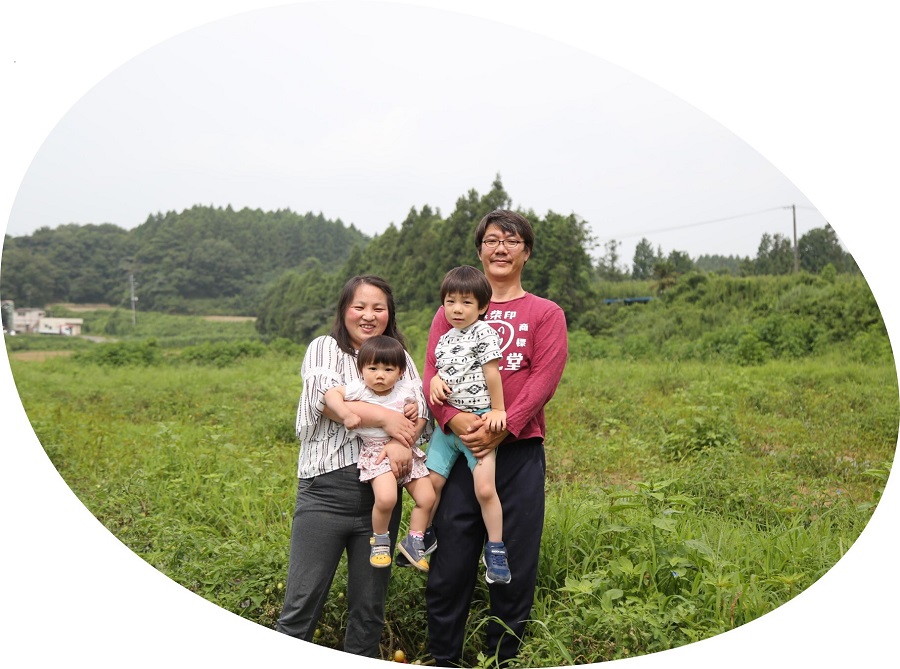 緑豊かな山々を背景にした広大な畑の中で、夫婦がそれぞれ幼い子どもを一人ずつ抱きかかえ、家族四人で仲睦まじく寄り添いながらカメラを見つめている桑原さん家族の写真