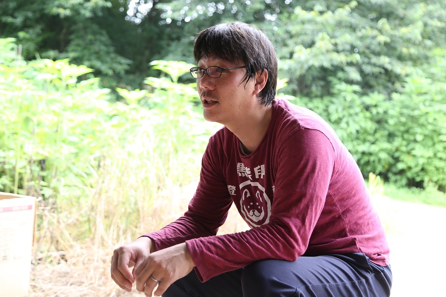 緑豊かな屋外で、眼鏡をかけた桑原さんが、屈みながら前方を見つめて穏やかに話しているインタビューの様子の写真