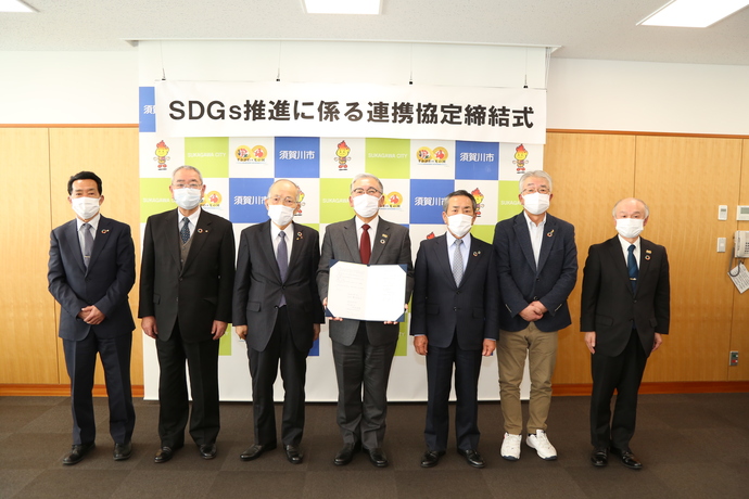 「SDGs推進に係る連携協定締結式」と書かれた横断幕を背景に、スーツやジャケット姿の男性7人がマスクを着用して一列に並び、中央の男性が協定書を広げて持って立っている記念写真