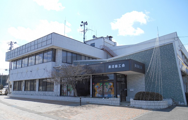 入口横の植栽に電飾が施され、白いタイル張りの壁面と大きな窓が特徴的な二階建ての建物の屋上に、アンテナや拡声器が設置されている長沼商工会の外観写真