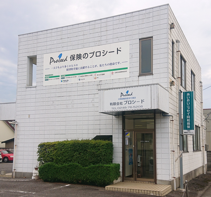 入り口や外壁に「保険のプロシード」と書かれた看板が設置され、入り口横に植栽が設けられた、白壁の二階建てビルの外観写真