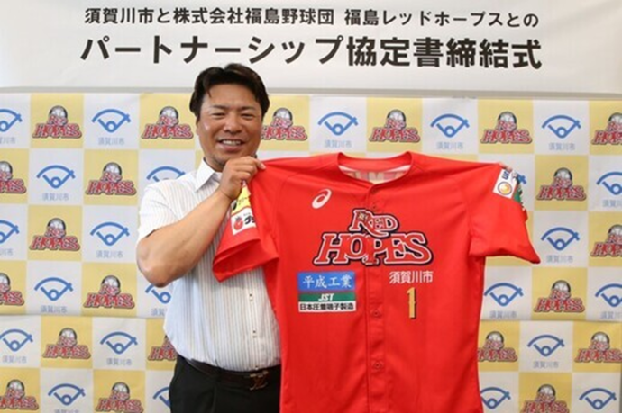 須賀川市と株式会社福島野球団福島レッドホープスによるパートナーシップ協定締結式において、笑顔の男性が「RED HOPES」と書かれた赤い野球のユニフォームを両手で広げて掲げてカメラに目線を送っている写真