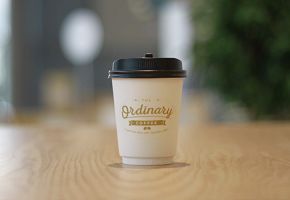 木製テーブルの中央に白い紙カップが置かれ、黒いプラスチック製の蓋が付いてカップには「The Ordinary Coffee Stand」という文字がゴールドで印刷されている写真