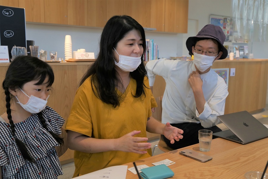 木製カウンターが背景にある明るい室内で、テーブルにノートパソコン、スマートフォン、ペン、メモ用紙、カード類、そして水の入ったグラスが置かれ、中央の女性は黄色の服を着て手を動かしながら説明し、左側にはチェック柄の服を着た女の子、右側には帽子をかぶり白いシャツを着た男性が座っている写真