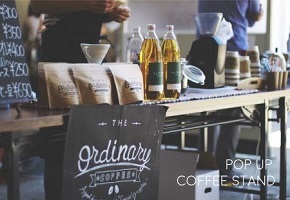 木製テーブルの上に、コーヒー豆の袋が複数並び、隣には透明なボトルに入った液体が2本置かれ、奥にはドリッパーやペーパーフィルターを使ったコーヒー抽出器具、紙カップが置かれている写真