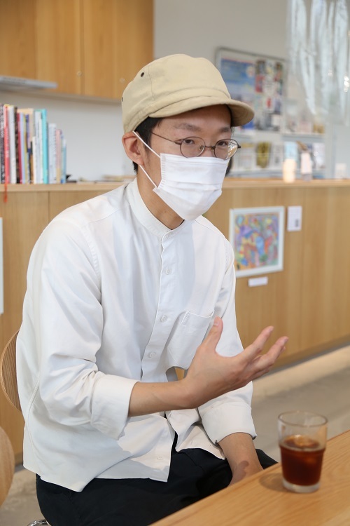 木製カウンターが背景にある明るい室内で、佑二さんが白いシャツとベージュのキャップを着用し、片手を前に出して会話や説明をしテーブルの上には氷入りのアイスコーヒー、背後のカウンターには本や装飾品、壁にはカラフルな絵が飾られている写真