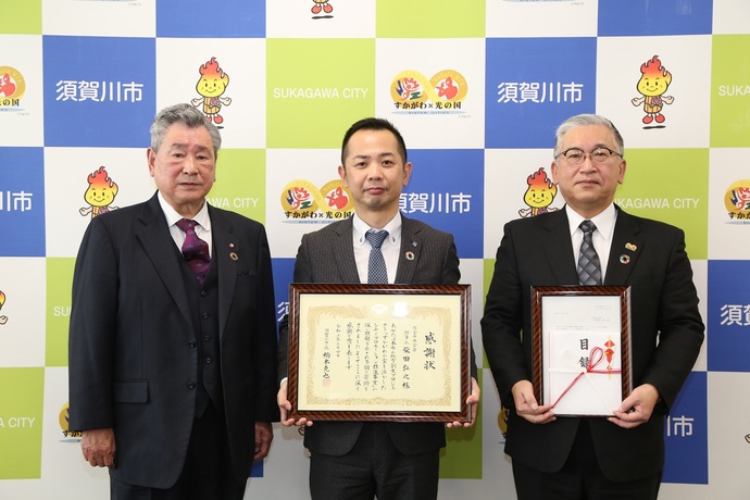 市のロゴが並ぶ背景の前で三人の人物が額装された感謝状や賞状を手に整然と並び立つ表彰式の様子を捉えた写真