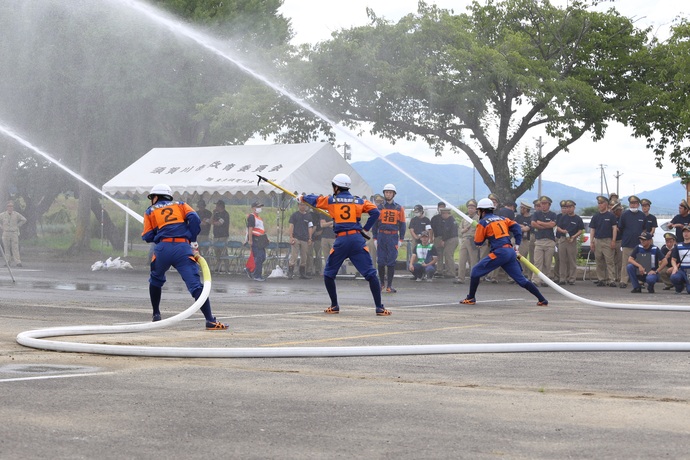 棒で斜め上を指している消防団員の指示に合わせ、両側に立った2人の消防団員が手に持ったホースから勢いよく放水している様子を、大勢の消防団員が見守っている様子の写真