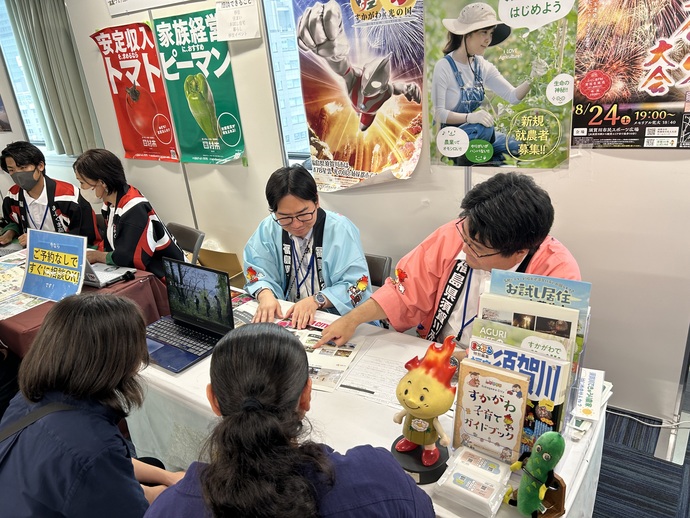 カラフルな法被を着た担当者が、相談ブースでパンフレットを指差しながら来場者に熱心に説明を行っている写真