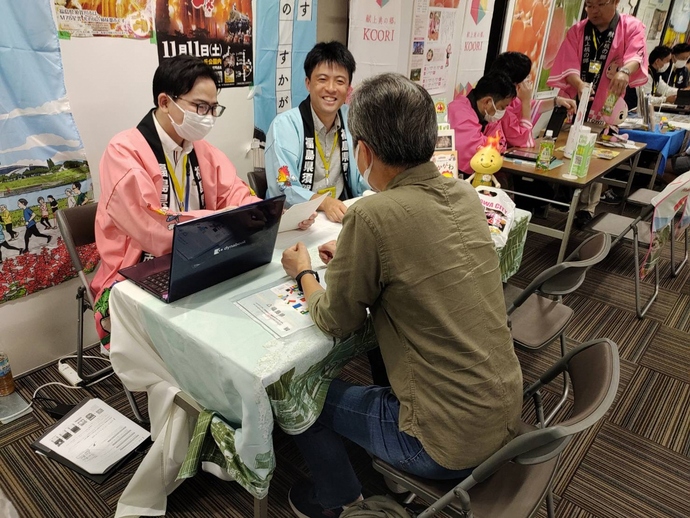 イベント会場のブースにて、ピンクや水色の法被を着たスタッフが、ノートパソコンや資料を使いながら、テーブル越しに座る男性客へ熱心に説明や相談対応を行っている写真