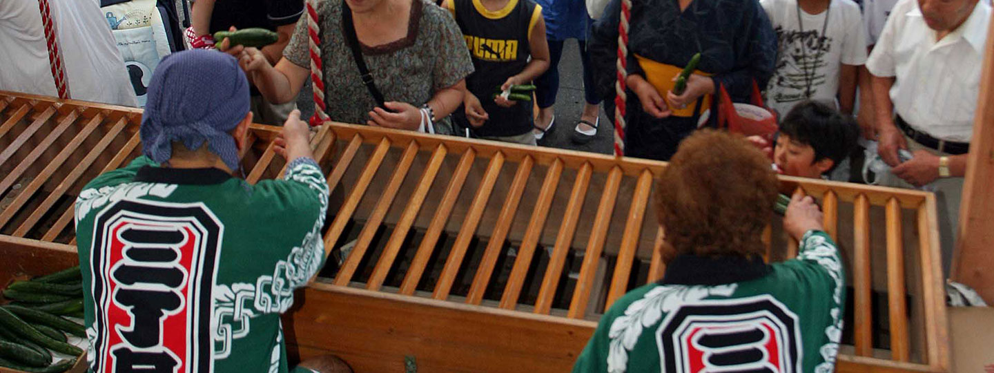 祭りの法被を着たスタッフが、木製の格子状の台越しに、並んでいる参拝客へキュウリを手渡している様子