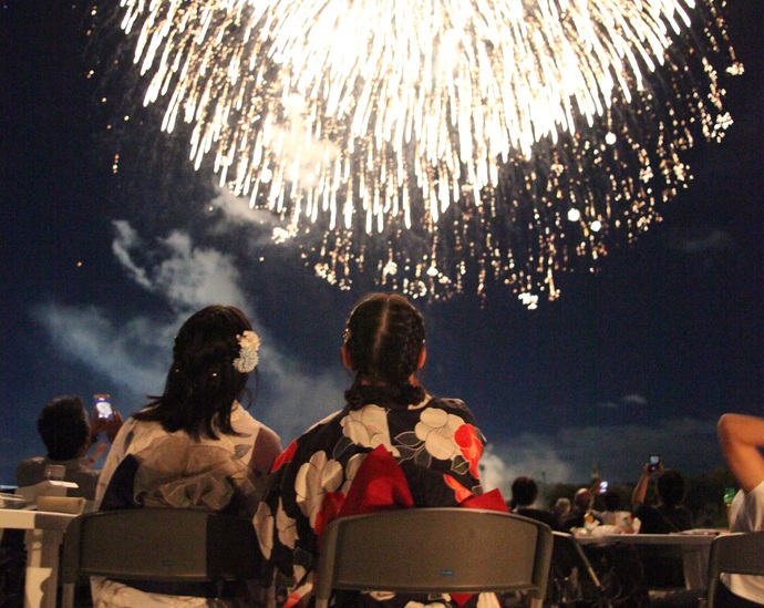 夜空を鮮やかに彩る巨大な打ち上げ花火を、椅子に座った浴衣姿の二人の女性が見上げている後ろ姿の写真