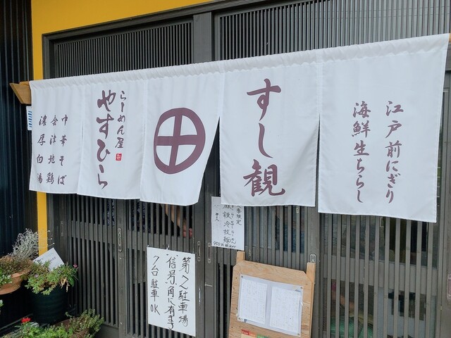 黒いサッシの引き戸が続く店舗の外観を捉えた写真で、中央には白地の暖簾が5枚かかっており、「すし観」の文字と丸に十字の家紋のような紋章、「らーめん屋 やすひら」の文字が染め抜かれている写真