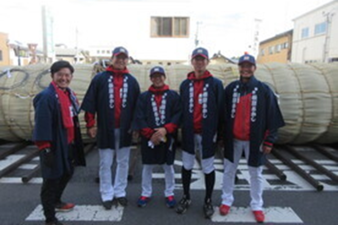横倒しに置かれた巨大な松明の前で赤いユニフォームの上に着た法被姿の5人が整列し笑顔を見せる祭りの様子の写真