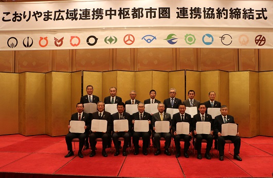 赤い絨毯が敷かれた壇上で、スーツ姿の人物たちが二列に並び、全員が白い書類を手に持って座っており、背後には金色の屏風が立てられ、上部には「こおりやま広域連携中枢都市圏 連携協約締結式」と書かれた横断幕と複数の自治体のシンボルマークが並んでいる式典の写真