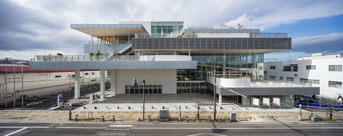 段状に重なるガラスと白い外壁が特徴的な現代的建築物が広い歩道沿いにそびえる写真